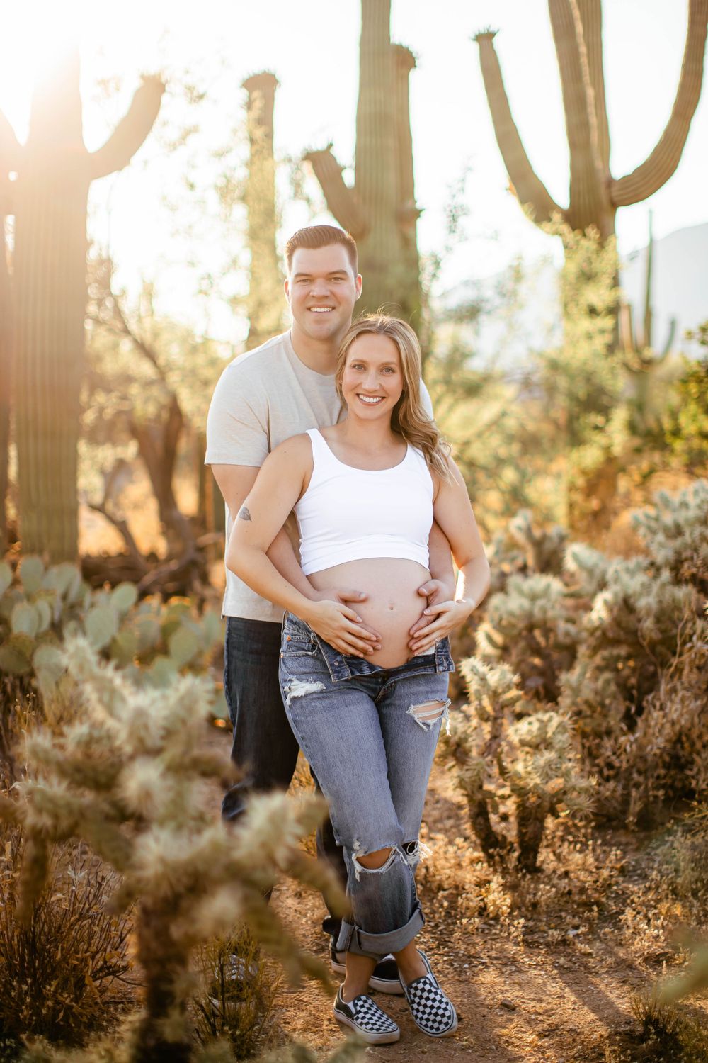 An expectant couple poses together among desert cacti during golden hour.