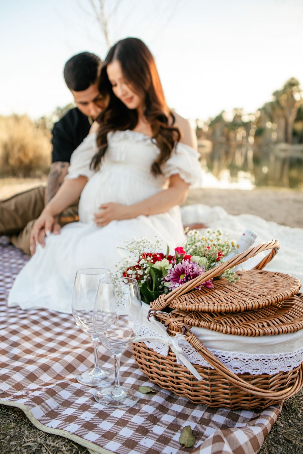 Romantic picnic setup with wicker basket, wine glasses and flowers on checkered blanket during golden hour sunset.