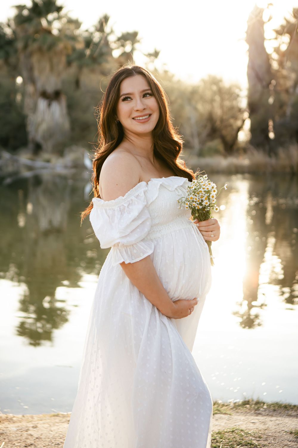 A woman in a flowing white dress stands by a serene lake at golden hour holding delicate baby's breath flowers.