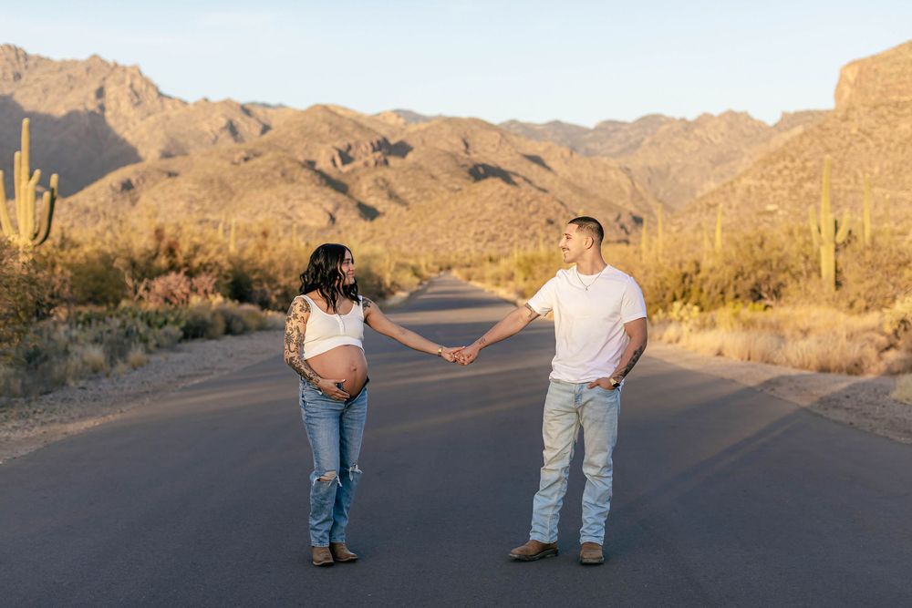 Couple holding hands while walking on desert road at sunset with saguaro cacti and mountains in background.