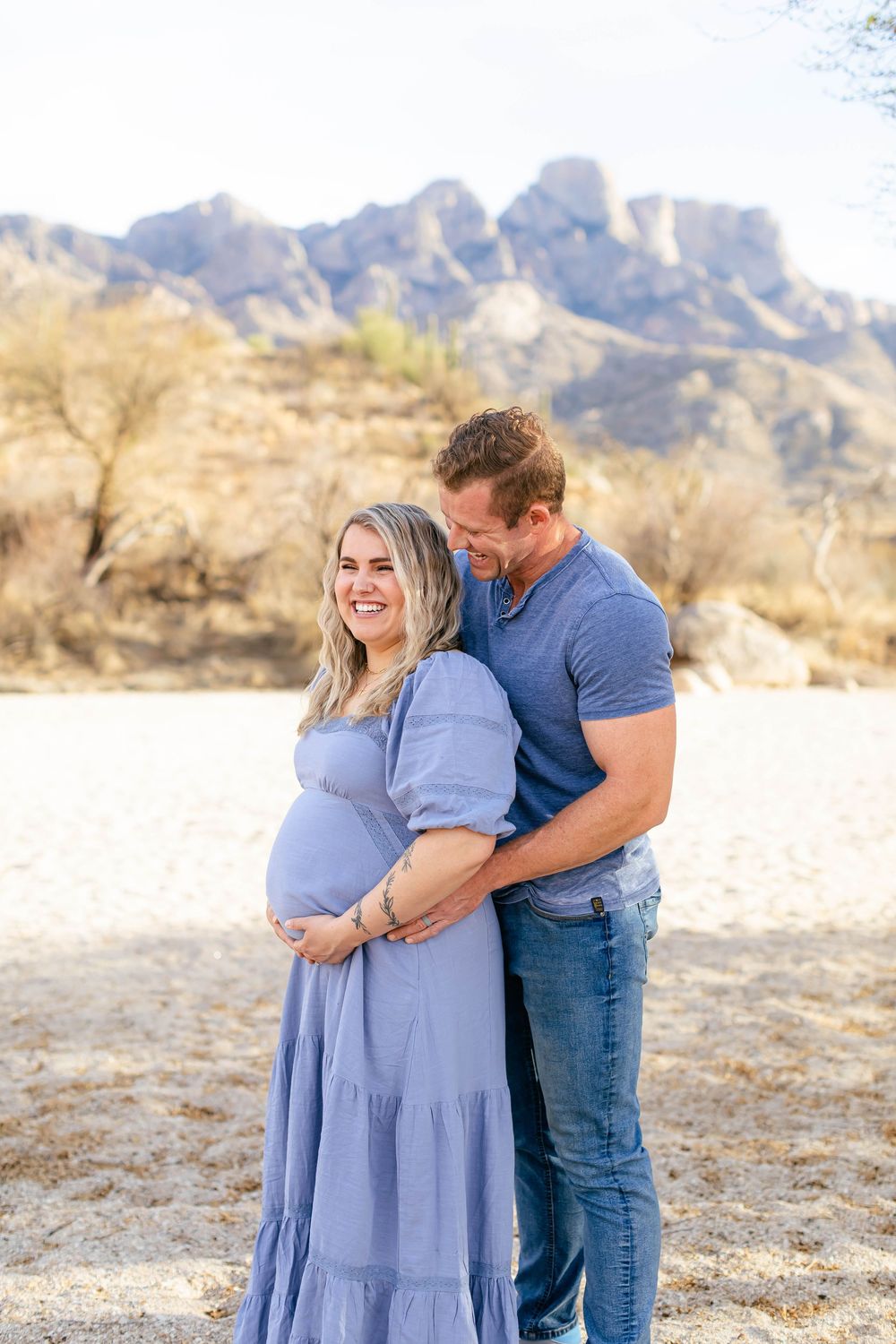 A couple embraces in a desert landscape with mountains in the background during a maternity photoshoot.