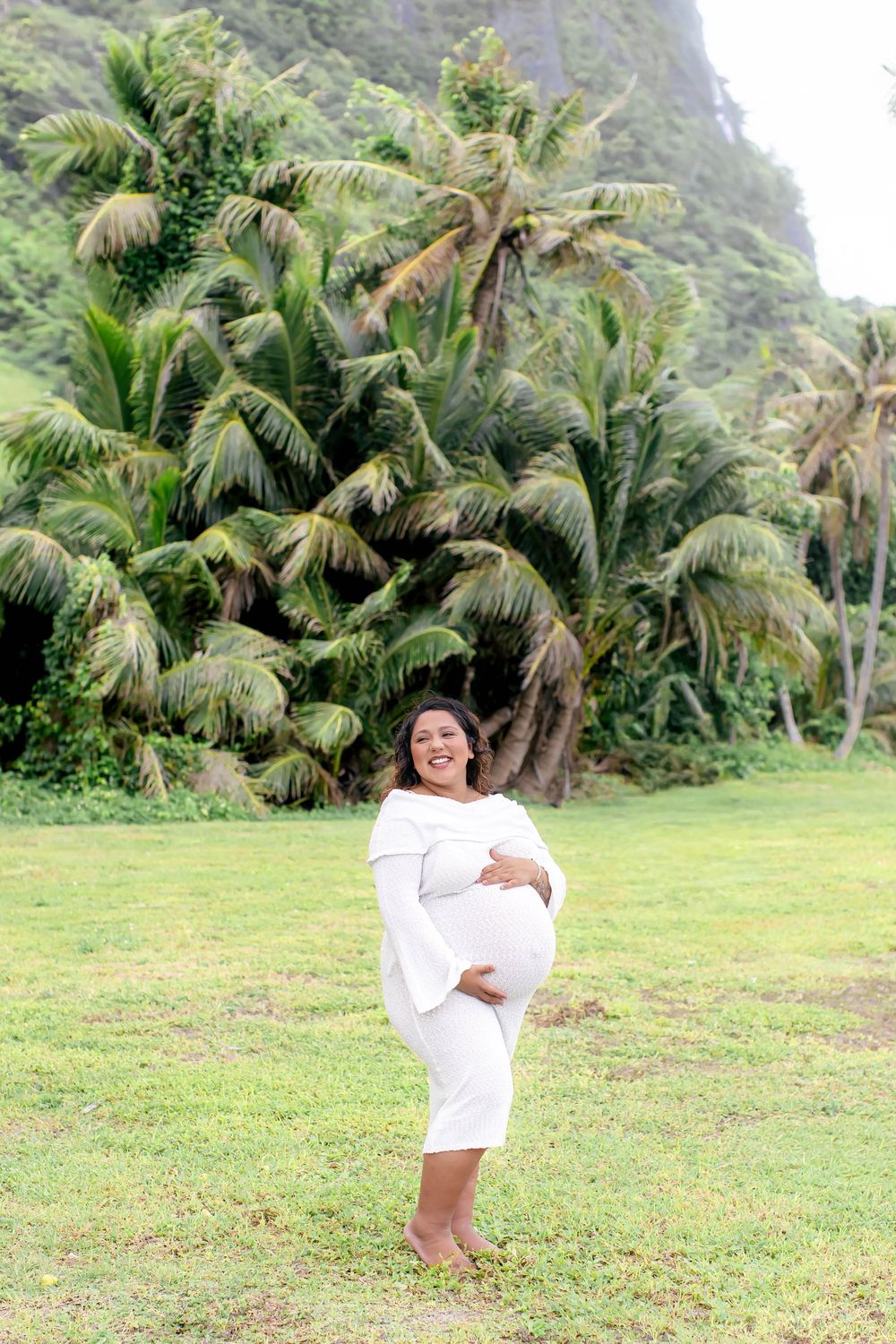 Smiling pregnant woman in white dress cradling belly outdoors with tropical palm trees and lush green mountains behind her.
