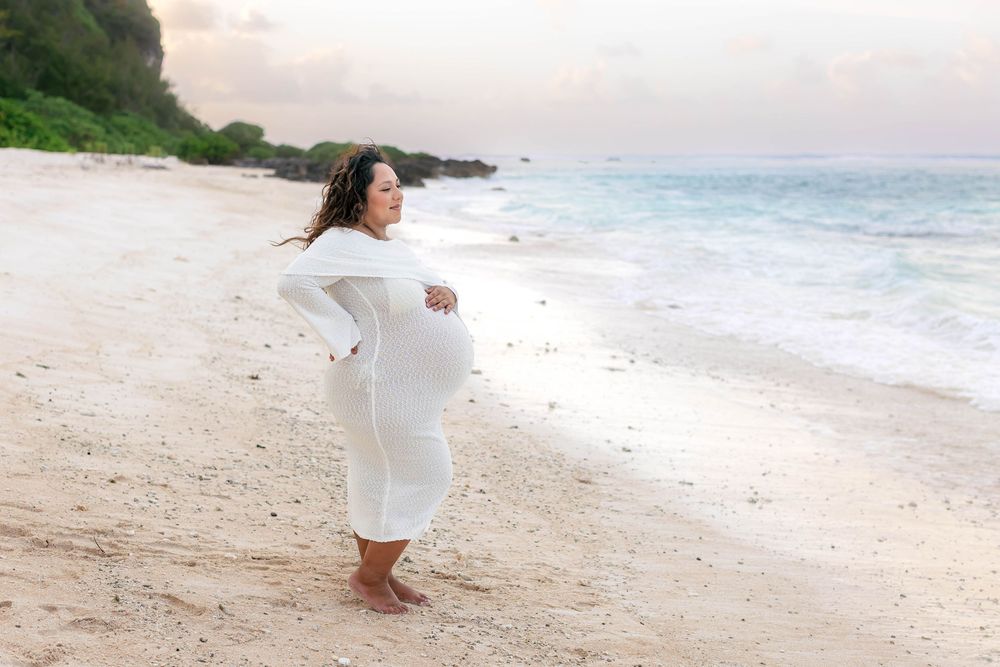 Pregnant woman in white dress standing on a sandy beach at sunset, cradling her baby bump near ocean waves.