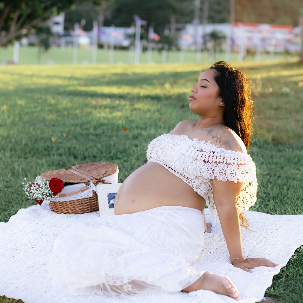 Pregnant woman in white lace outfit relaxing on blanket in park with picnic basket during golden hour.