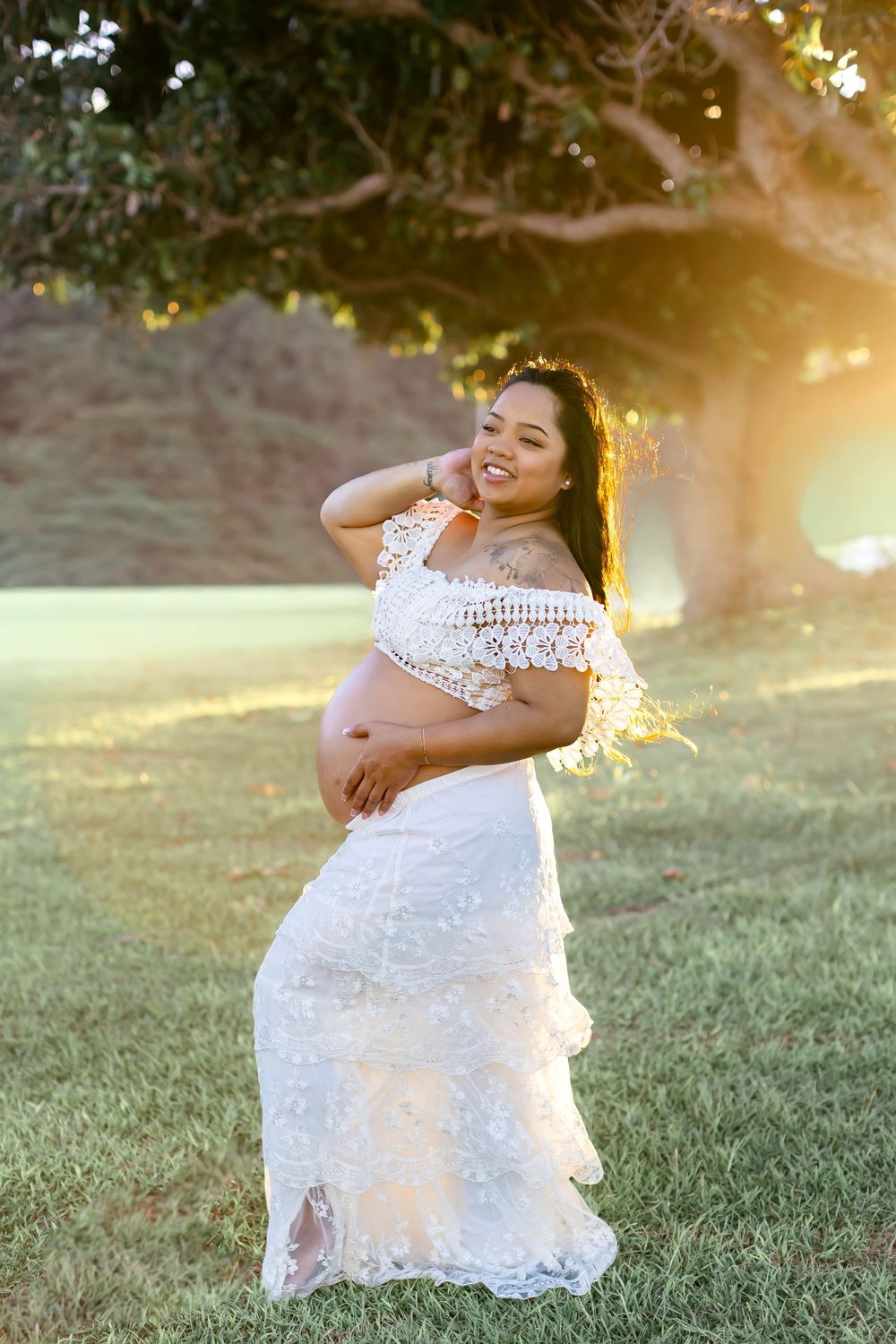 Pregnant woman in white lace crop top and skirt cradling baby bump outdoors at golden hour near a tree.