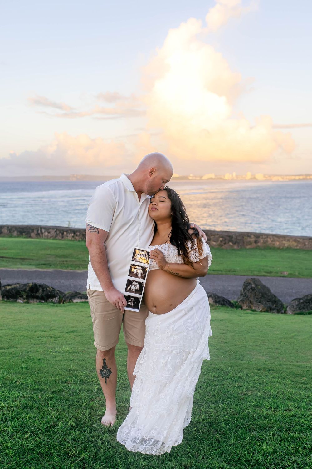 Couple sharing a tender maternity moment at sunset by the ocean, woman in white lace dress holding ultrasound photos.