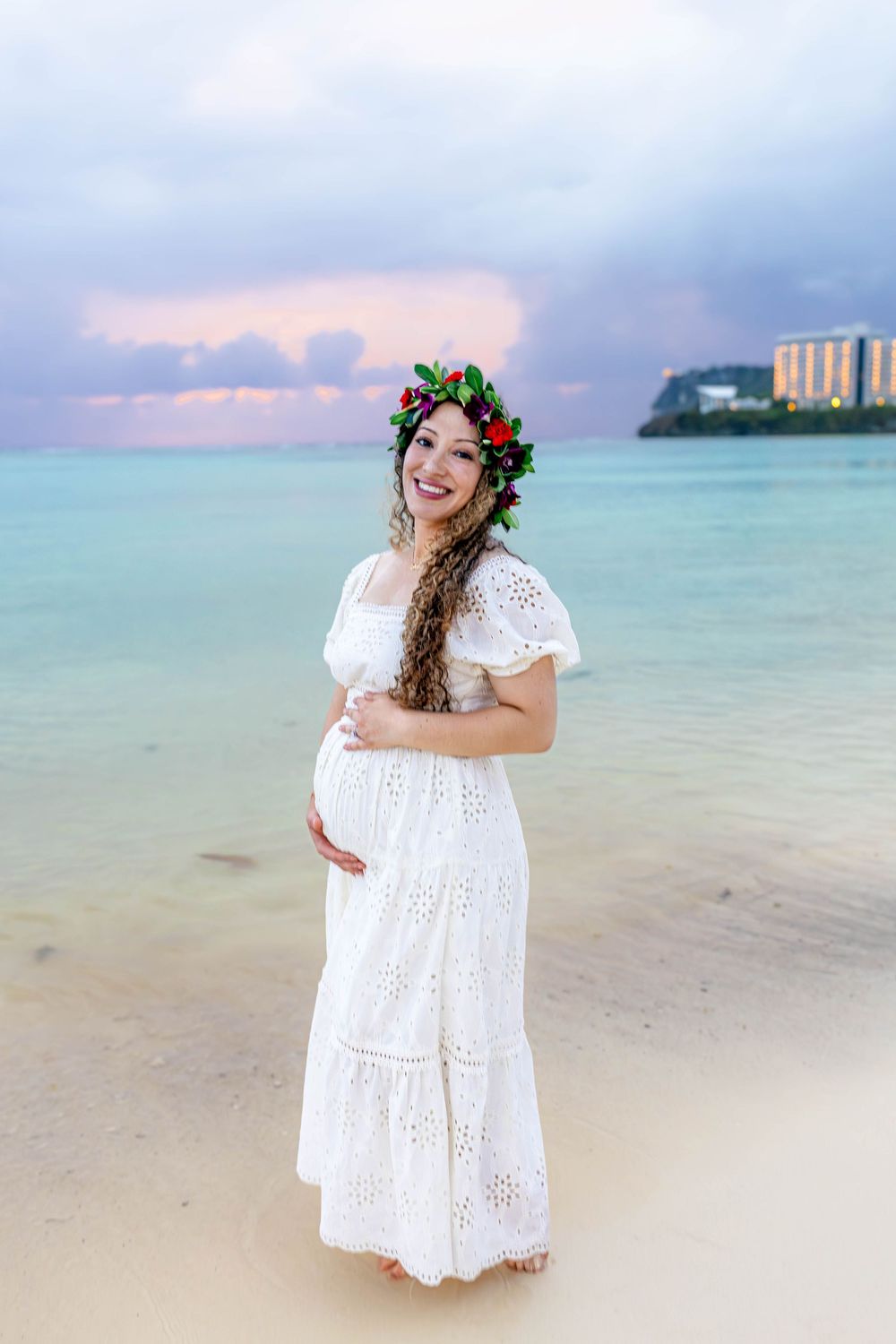 Pregnant woman in white eyelet dress and floral crown smiling on tropical beach at sunset.
