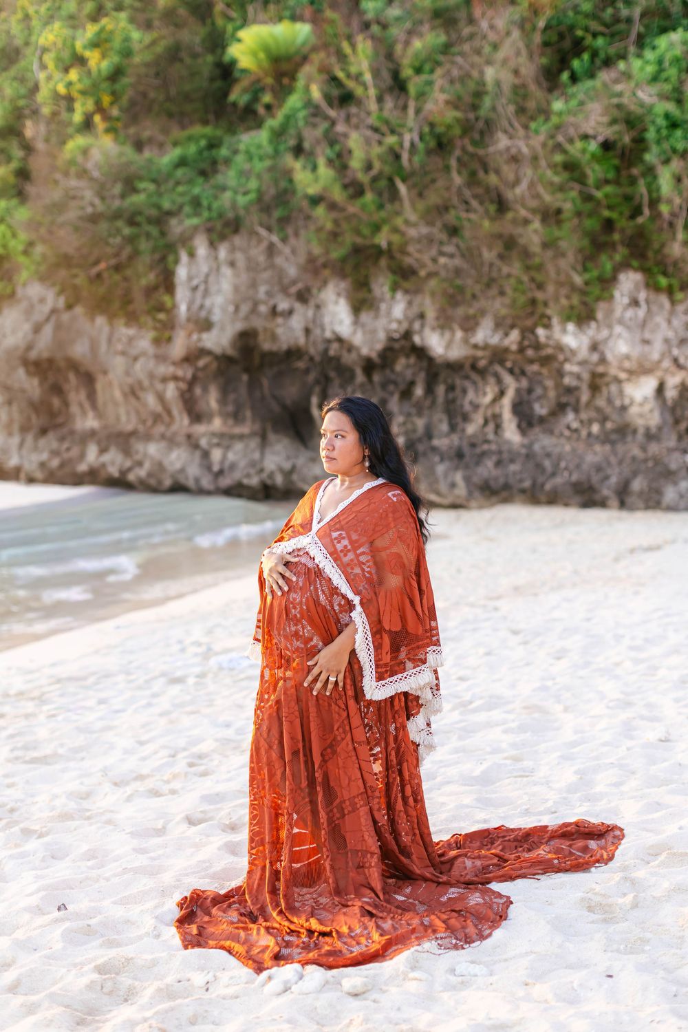Pregnant woman in flowing rust-orange gown poses on white sand beach with rocky cliffs and lush greenery behind her.