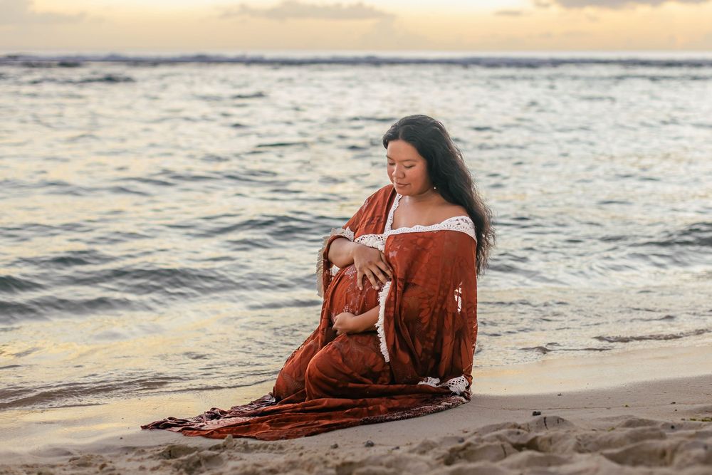 A mother sits on a sandy beach at sunset, cradling and breastfeeding her newborn wrapped in a flowing rust-colored fabric.