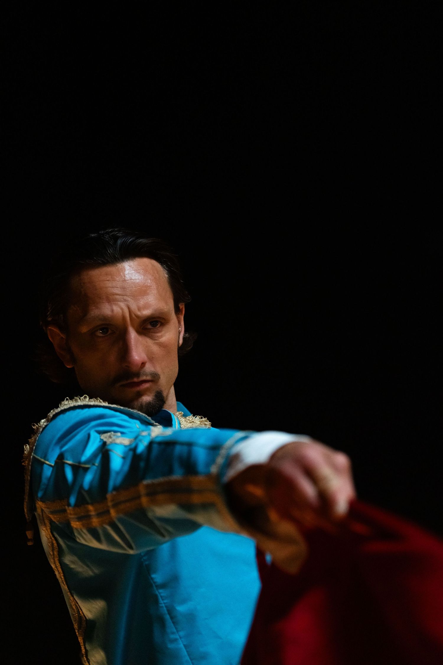 Dramatic portrait of performer in blue costume holding red fabric against dark background.