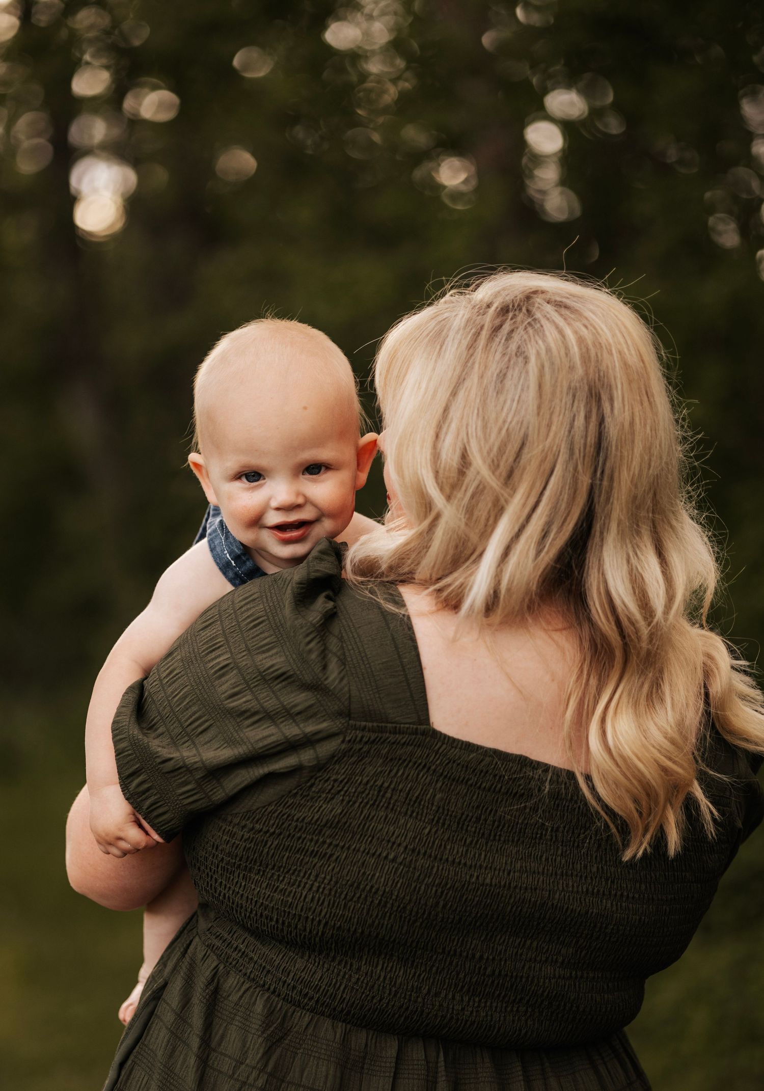 A baby smiles over the shoulder of someone in a dark dress against a bokeh forest background.