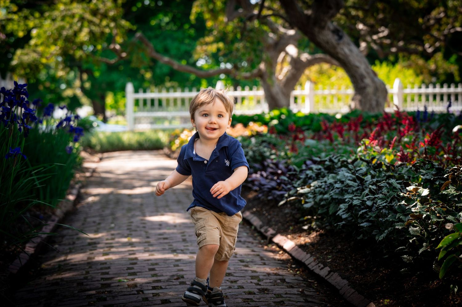 A young person runs joyfully along a brick path in a garden with purple flowers and white fence.
