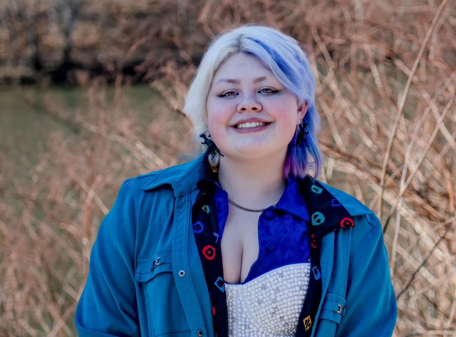 A person with blue hair wearing a denim jacket and white top poses outdoors against a natural background.