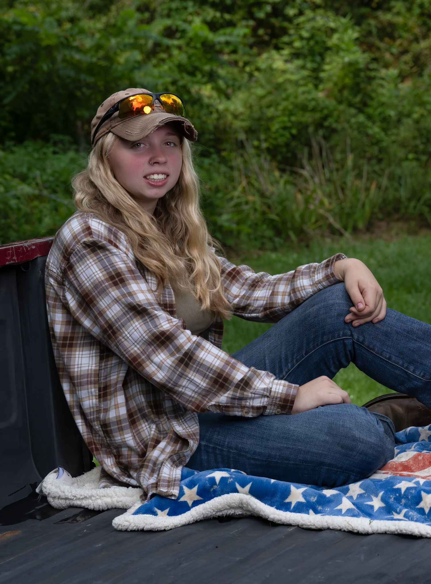 Person relaxing on American flag blanket wearing plaid shirt and baseball cap outdoors.