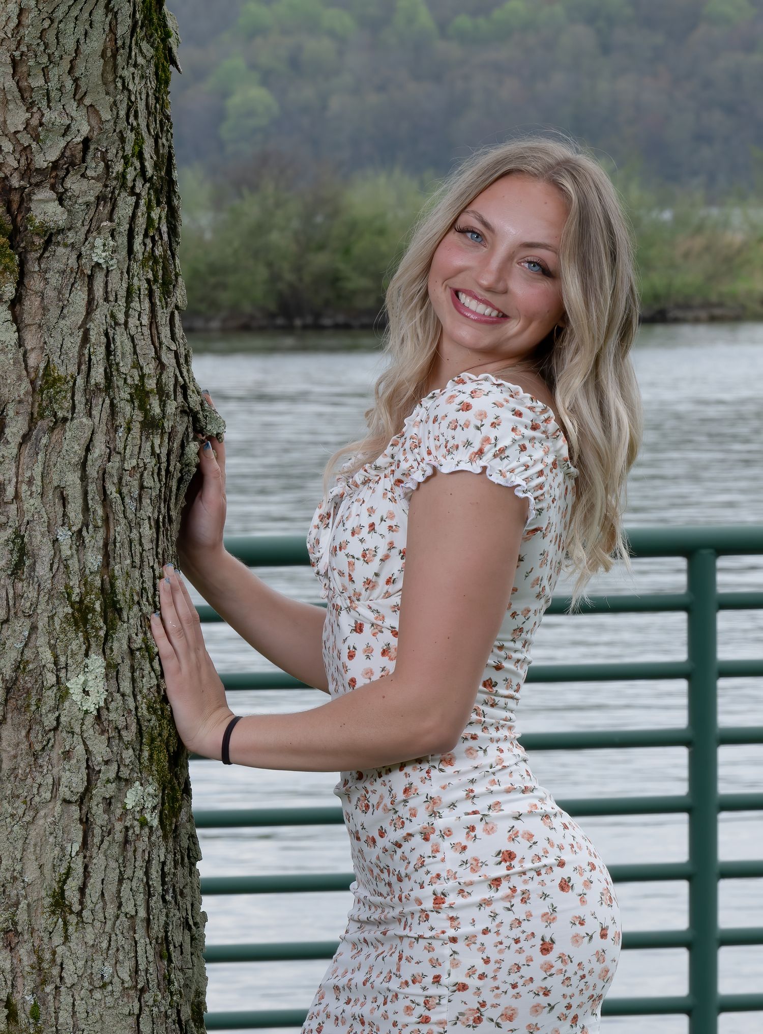 Person in floral dress poses by railing near water and trees in outdoor setting.