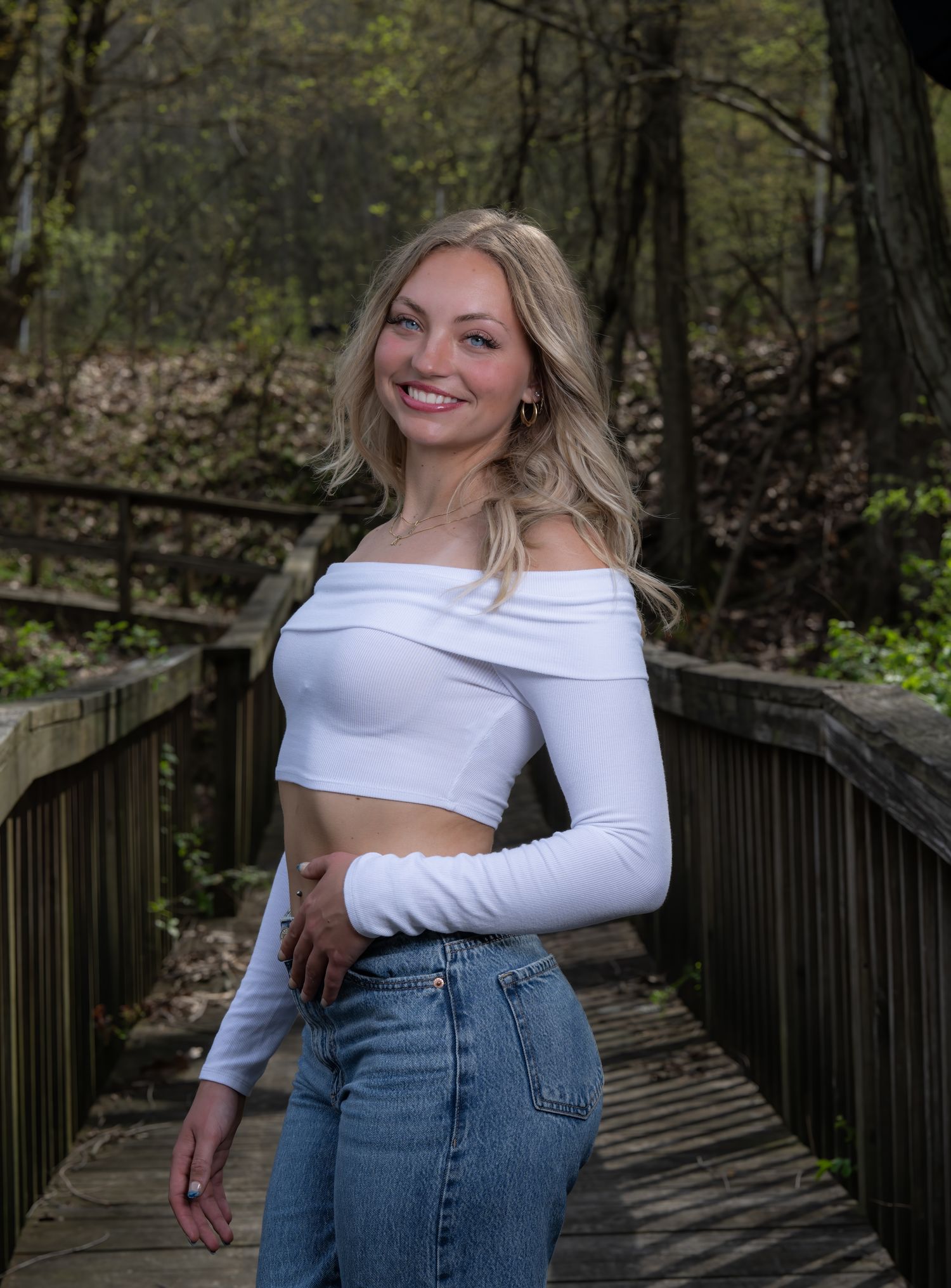 Portrait of person in white crop top and jeans on wooden bridge in forest setting.