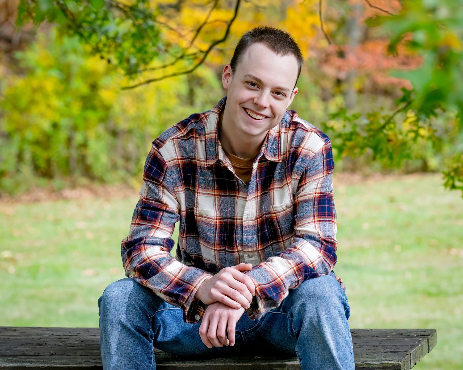 A casual outdoor portrait of a person in a plaid flannel shirt sitting on a bench surrounded by autumn foliage.