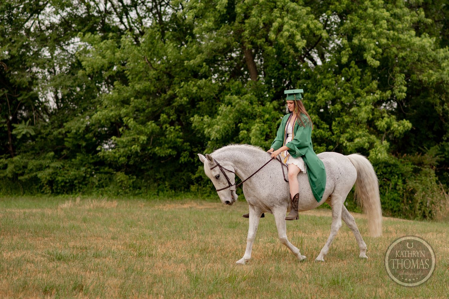 Graduate rides white horse through green field wearing traditional graduation attire.
