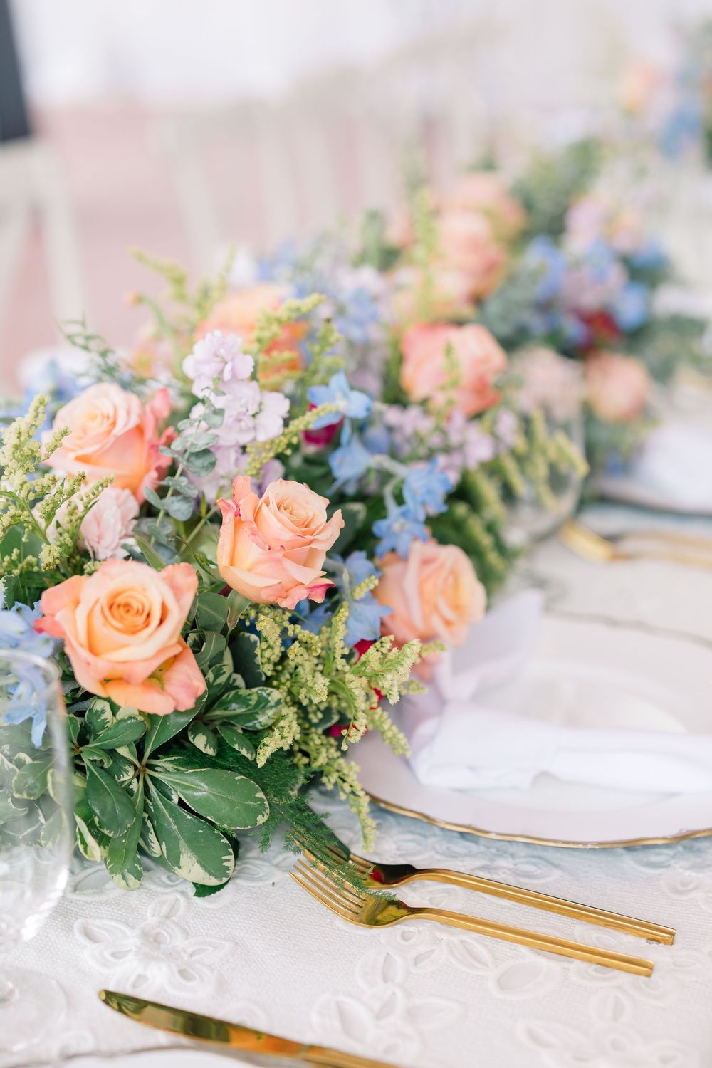 Elegant table setting with peach roses and blue flowers arranged with gold cutlery and white linens.