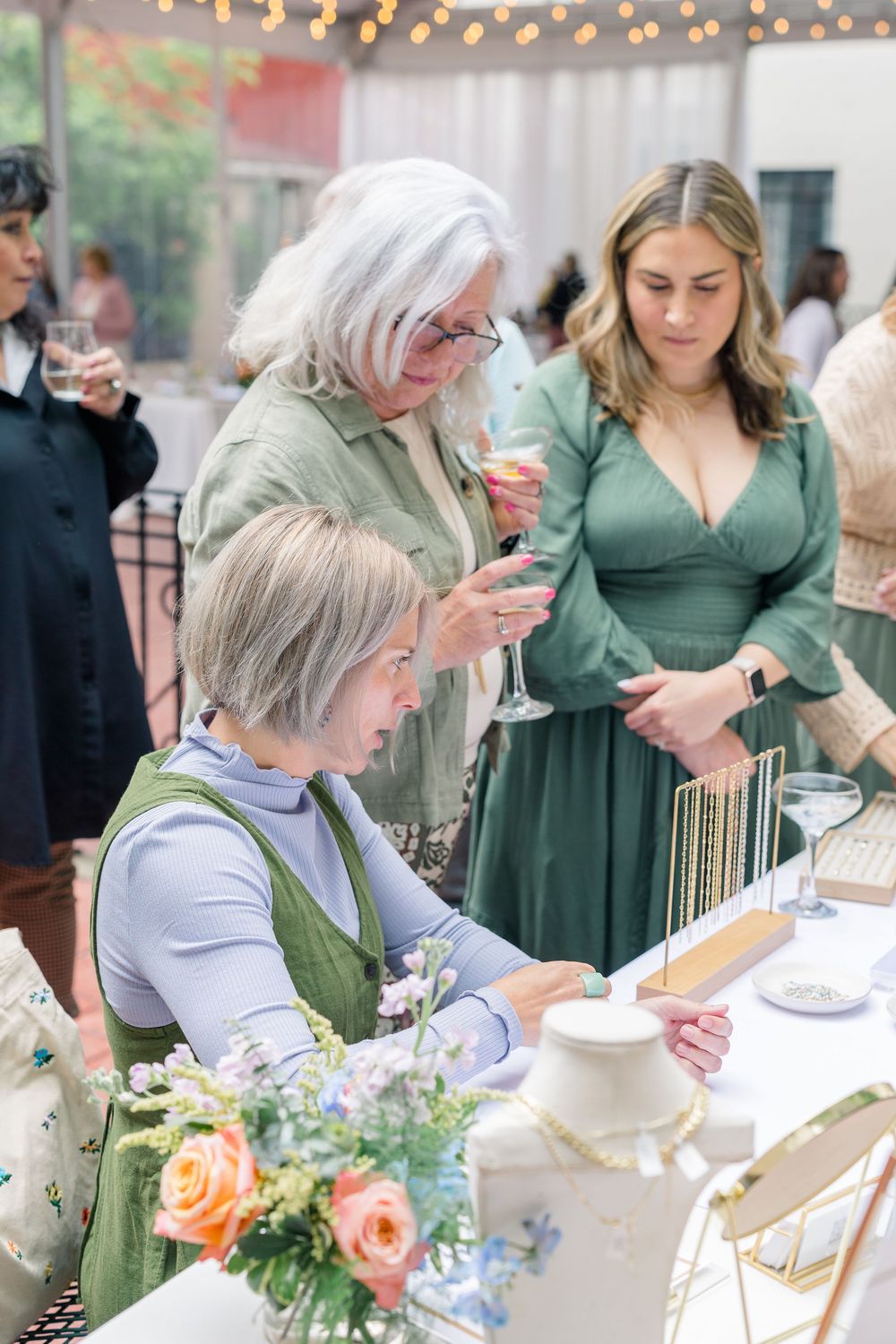 People gathered at an elegant outdoor event with string lights, looking at items on a table decorated with floral arrangements.