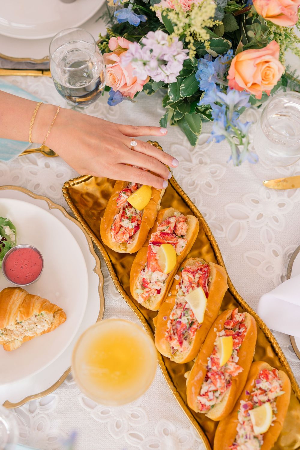 Overhead view of lobster rolls and wine glasses arranged on an elegant table setting with coral flowers and greenery.
