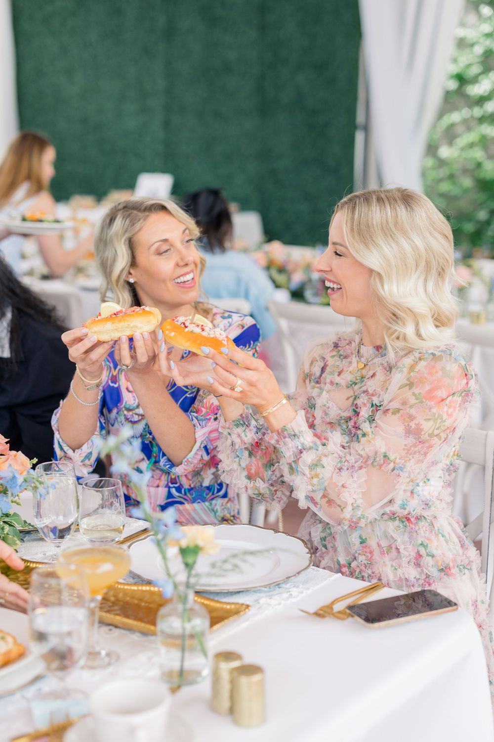 Two people in floral dresses share laughter while enjoying a meal at an elegant outdoor dining table.
