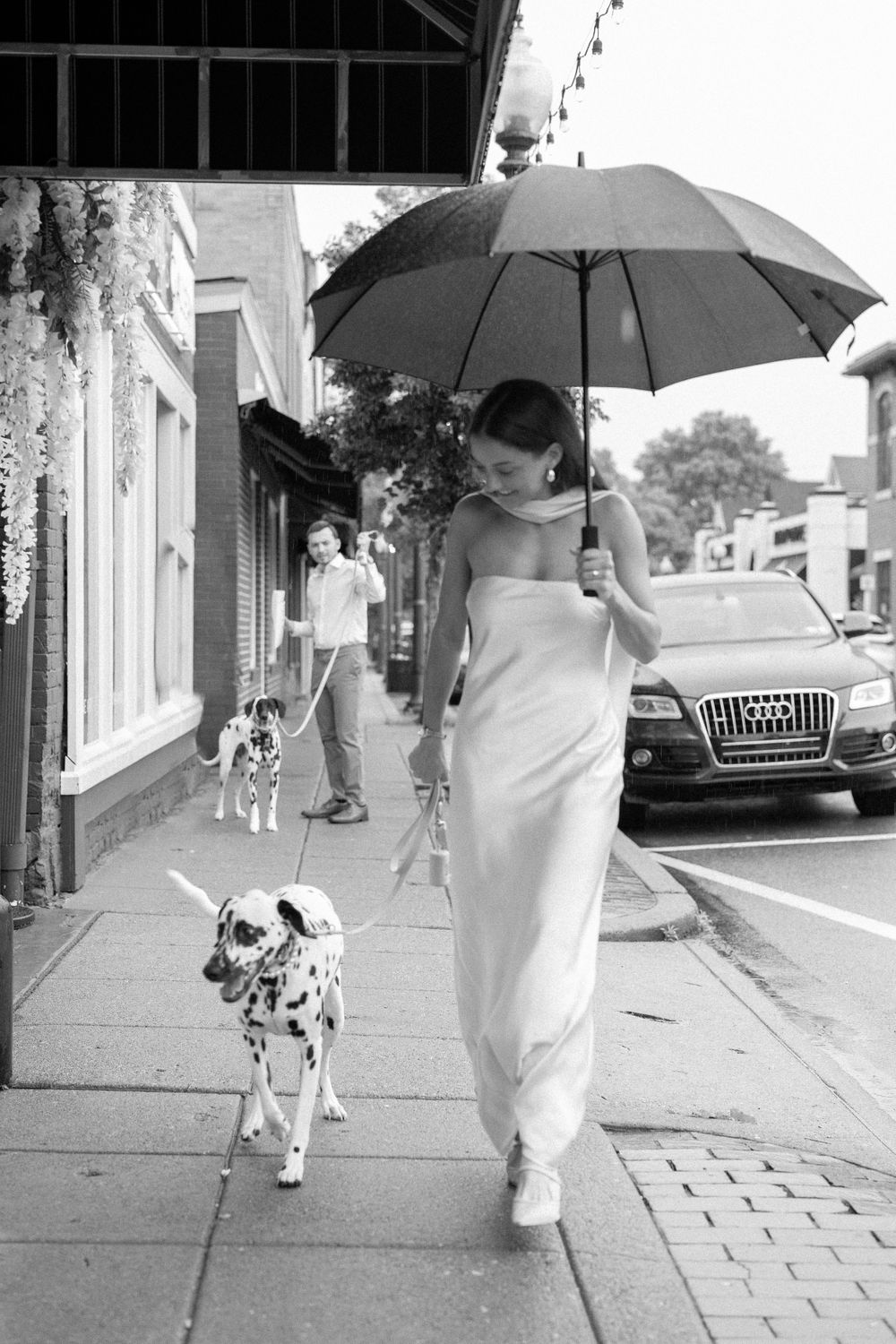 Person in white dress walking Dalmatian dog down urban sidewalk with umbrella.