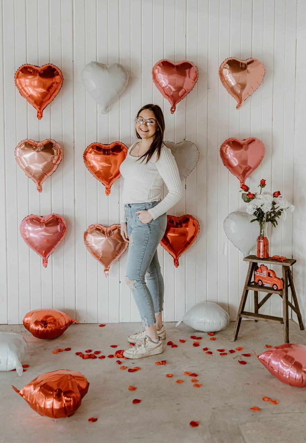 Woman in white sweater posing with heart-shaped balloons against white paneled wall.