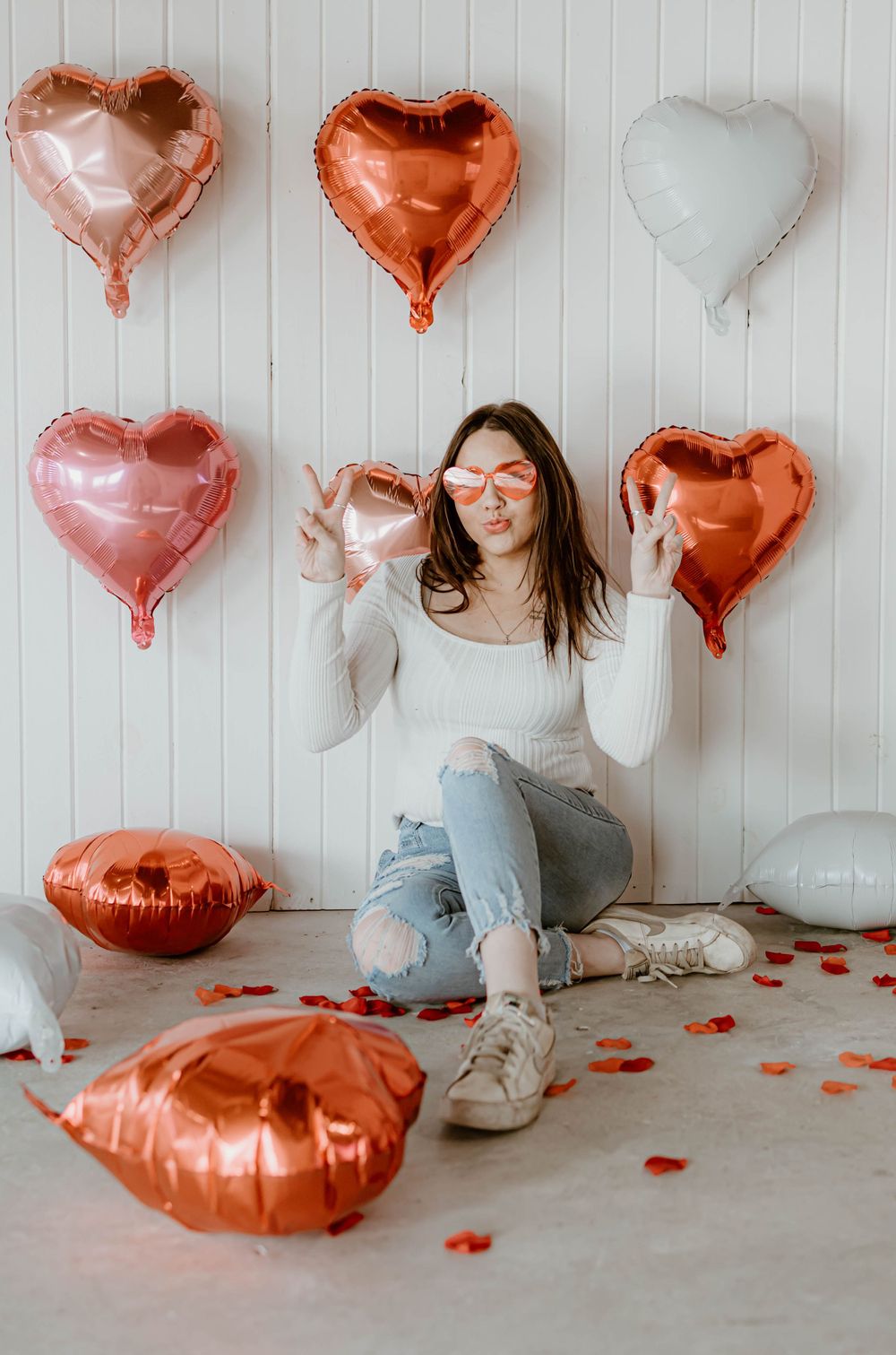 Playful Valentine's photoshoot featuring metallic heart balloons and scattered rose petals on floor.