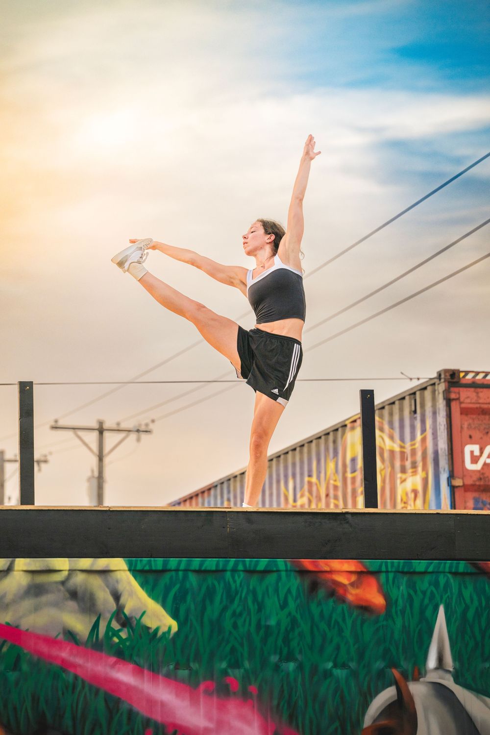 Gymnast performs graceful splits pose on outdoor stage with sunset sky and urban setting in background.