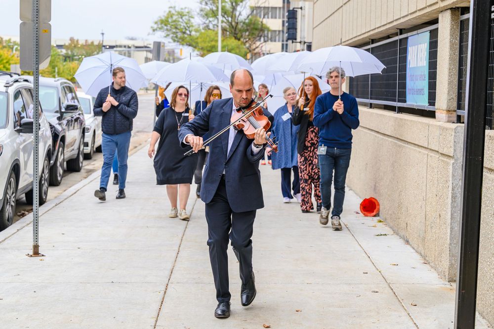 Wedding party members walk down a city sidewalk carrying white umbrellas on an overcast day.