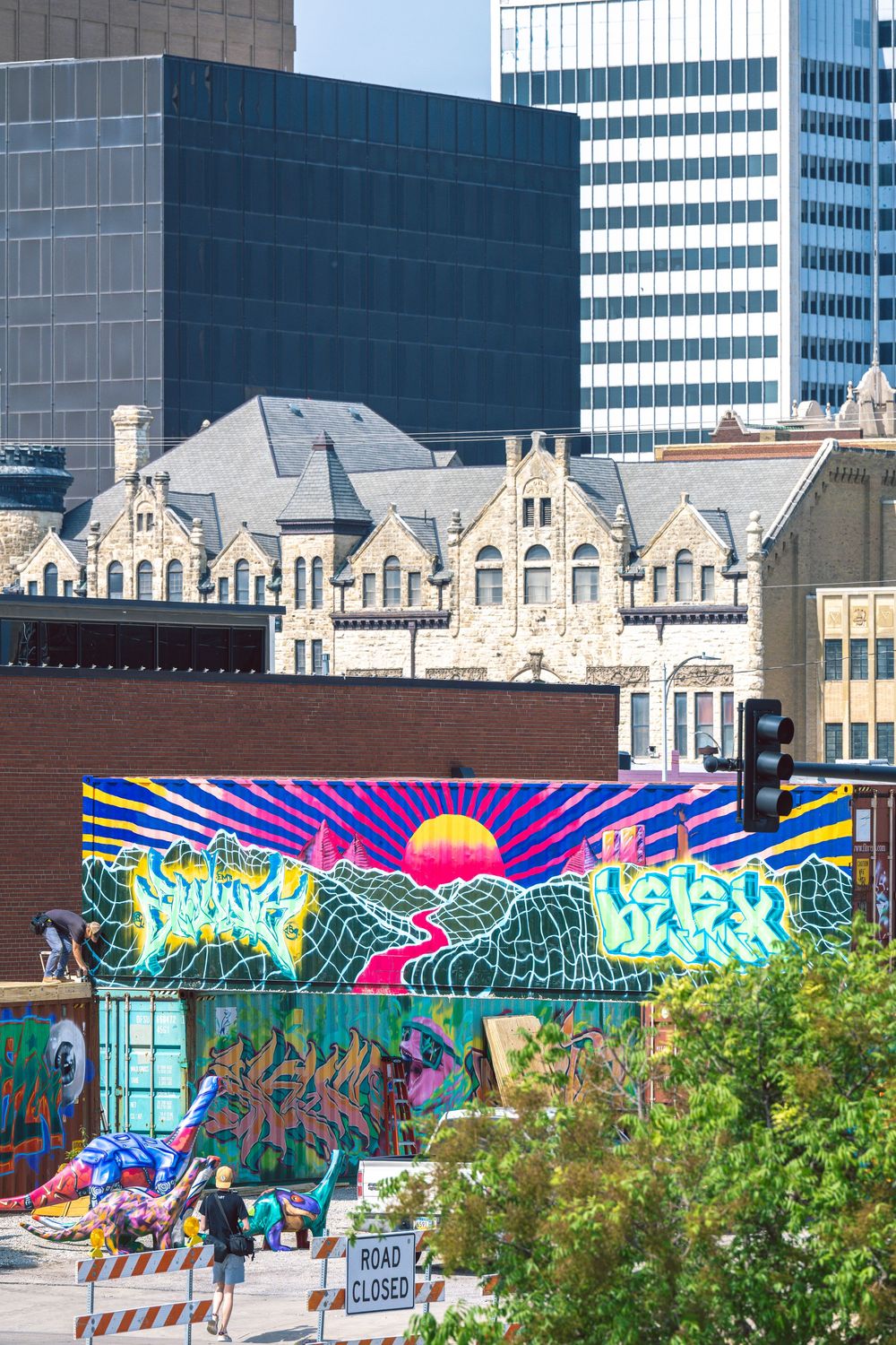 Colorful urban street art murals decorate walls along a city street with modern buildings and historic architecture in background.