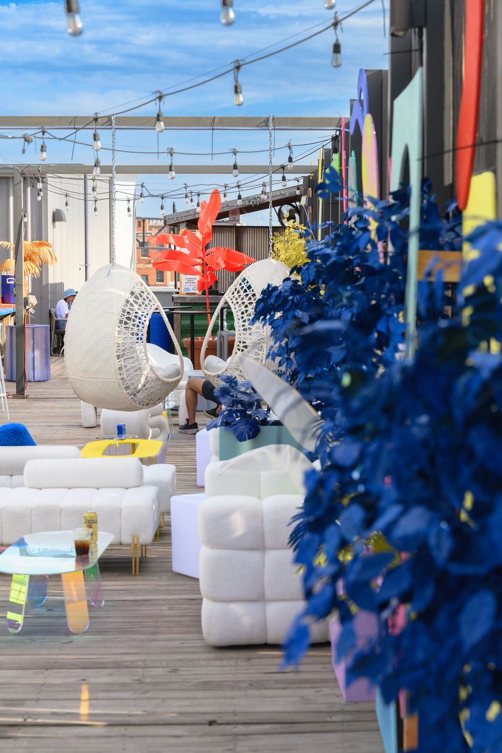 Modern rooftop terrace with hanging lights, white wicker chairs, and vibrant blue decorative elements on a wooden deck.