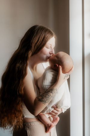 Tender moment between mother with long hair and infant by window in natural light.
