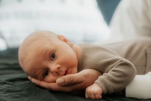 Newborn baby in knit sweater rests head on hand against dark fabric.