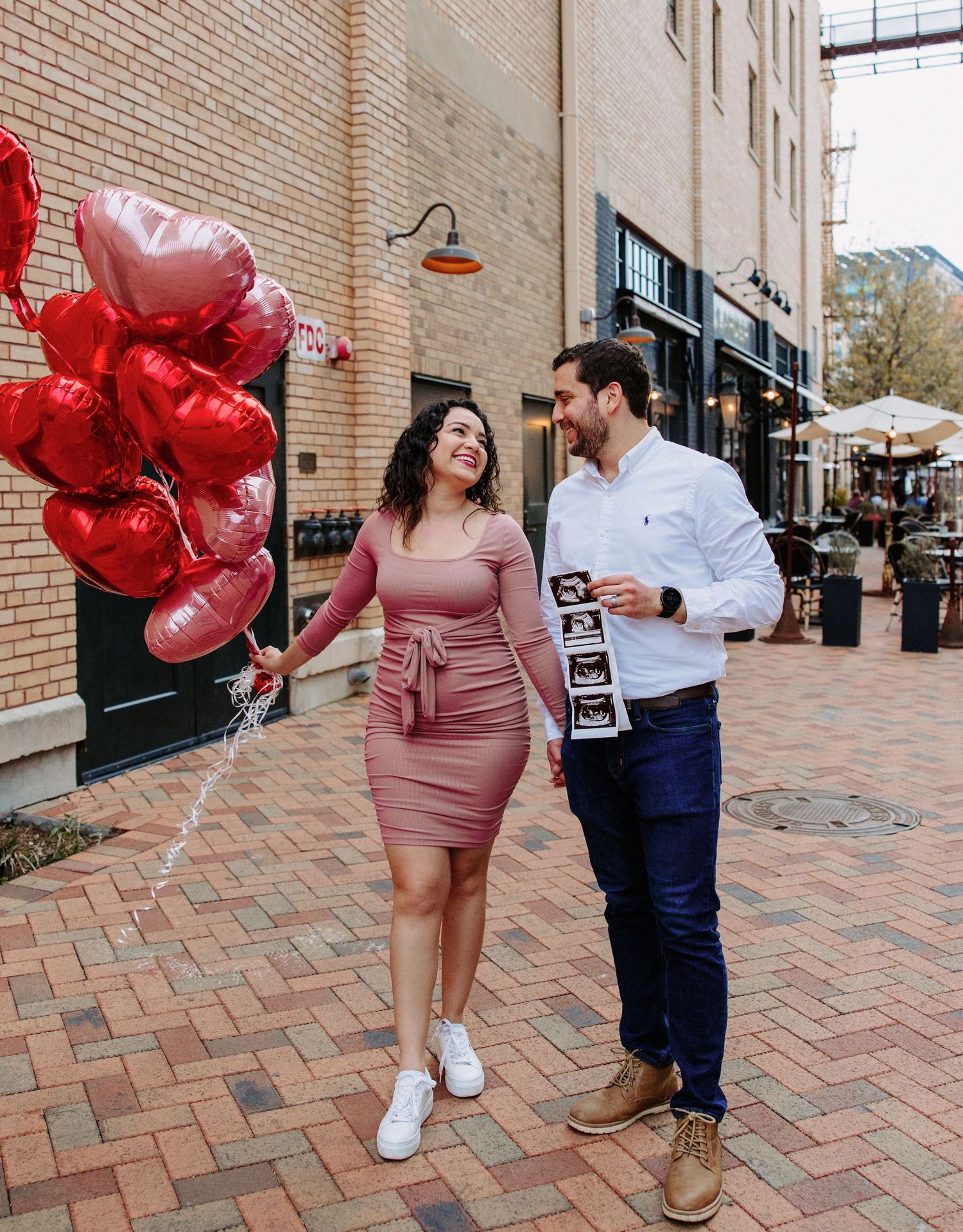 San Antonio Maternity Photography. Couple sharing a moment on brick sidewalk with red heart balloons against urban storefront.