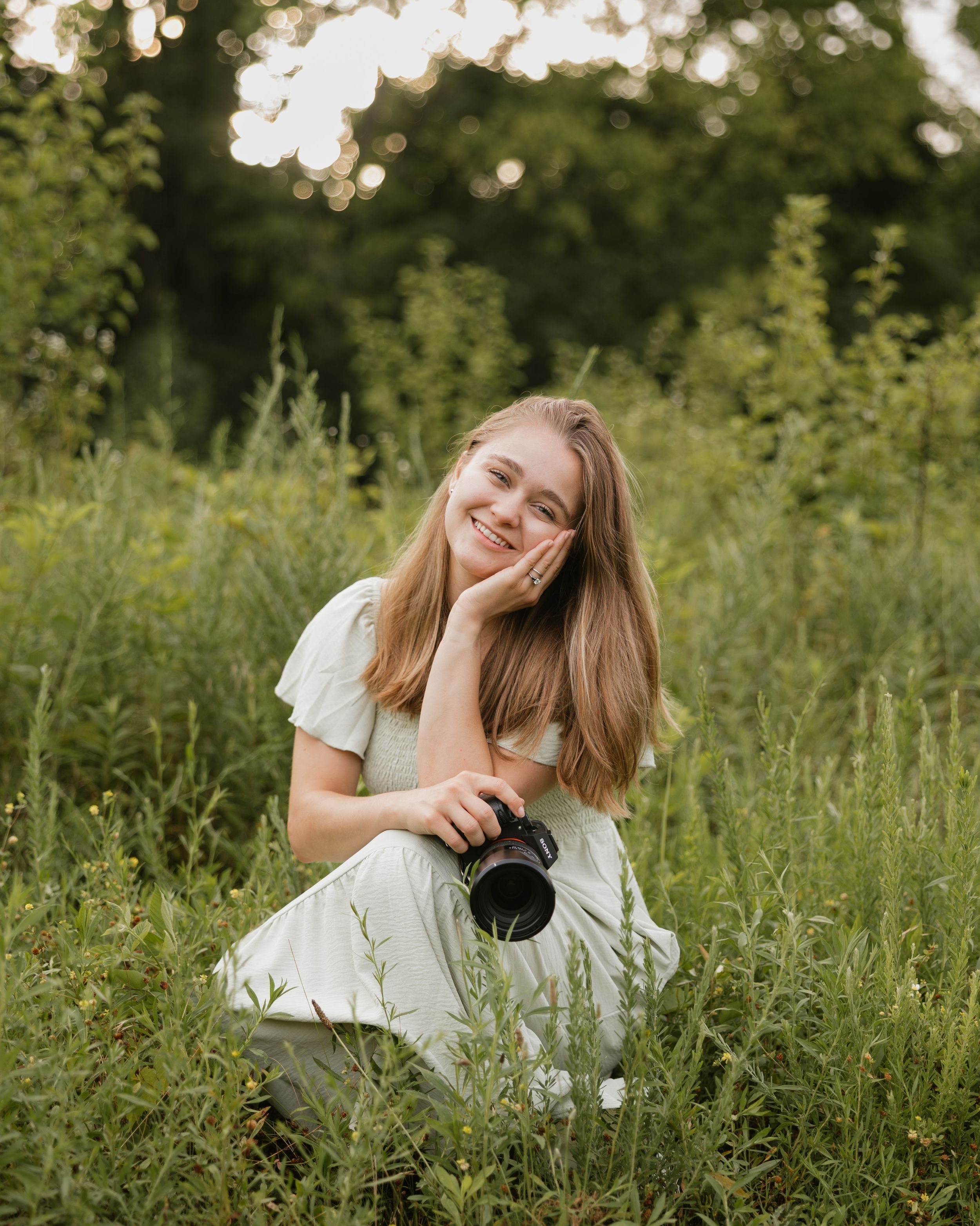 Woman in casual white sweater and blue jeans inspects camera while seated on white chair against neutral background.