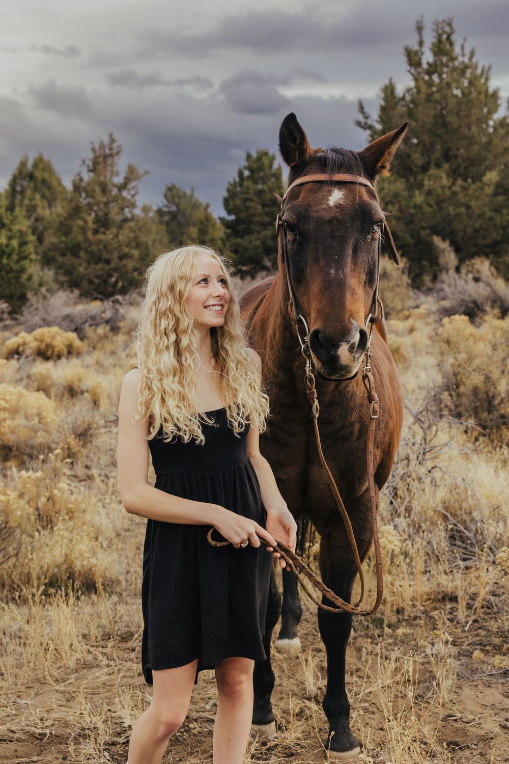 Person in black dress holding reins of brown horse in desert setting.