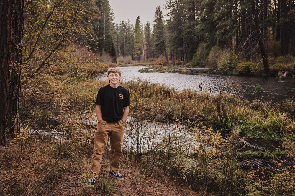 A casual portrait in front of a scenic river with pine trees in autumn.