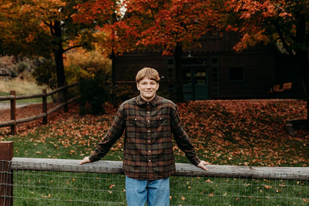 Someone wearing a plaid flannel shirt stands by a wooden fence surrounded by vibrant autumn foliage and fallen leaves.