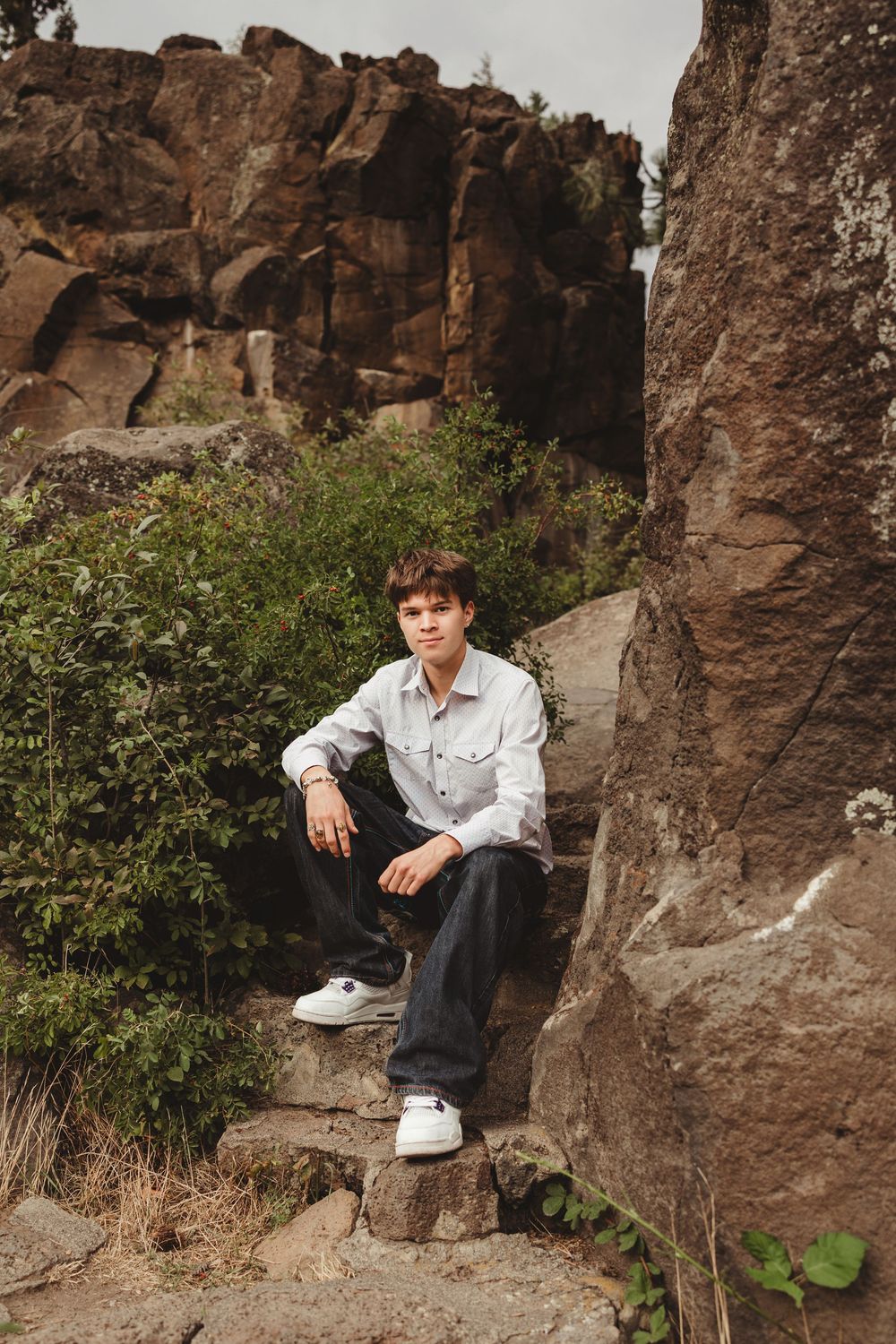 A figure sits on rugged brown rocks surrounded by green foliage in an outdoor setting.