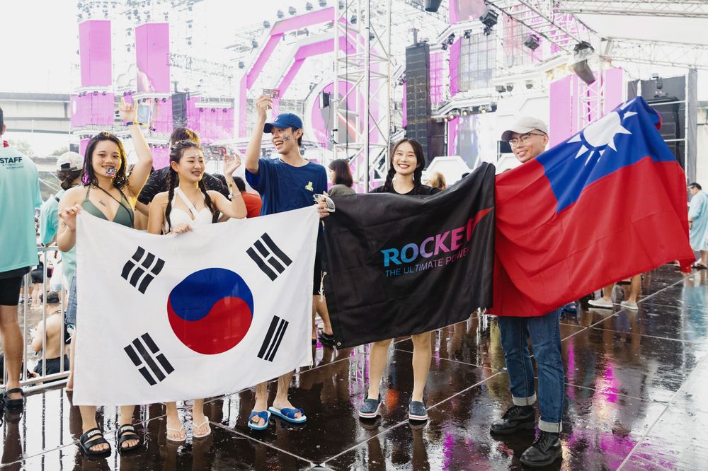 Festival attendees holding South Korean flag, Rocket banner, and Taiwan flag at outdoor event.