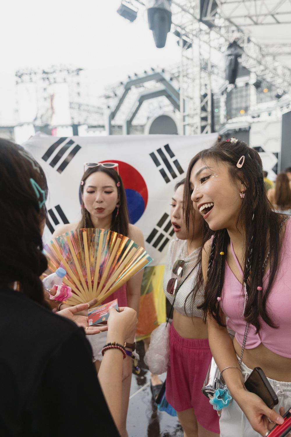 Young women in colorful festival outfits laughing near South Korean flag at music event.