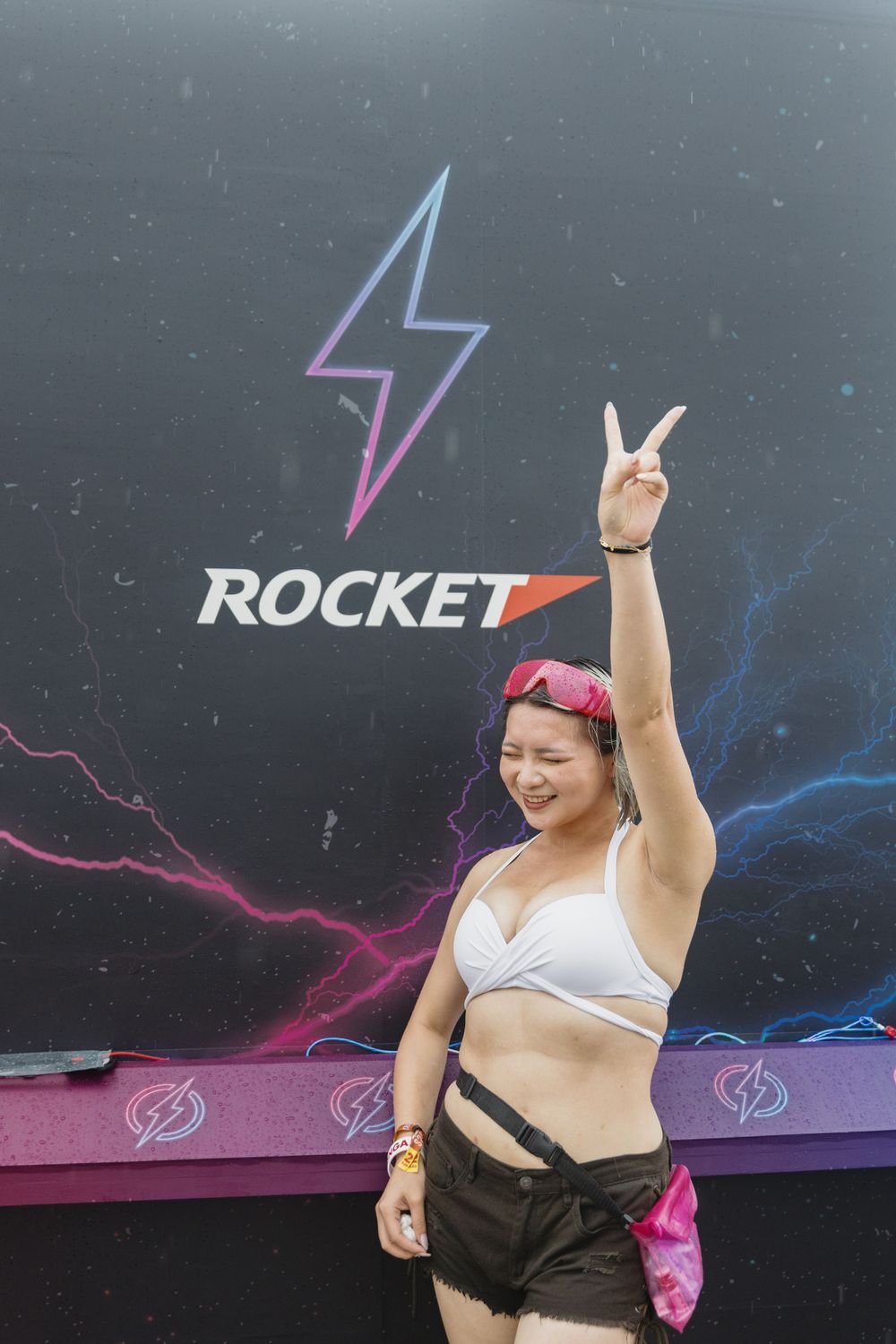 Woman in white bikini top flashing peace sign in front of Rocket brand backdrop.