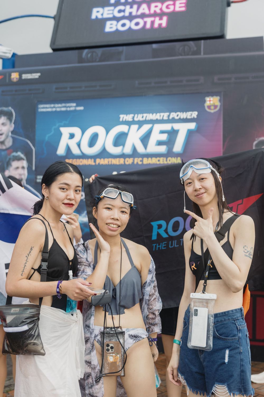 Three women posing in front of Rocket energy drink recharge booth at outdoor festival.