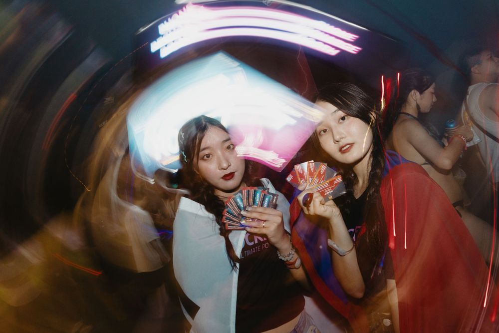 Two young women posing with colorful cards under neon lights at lively nightclub party.