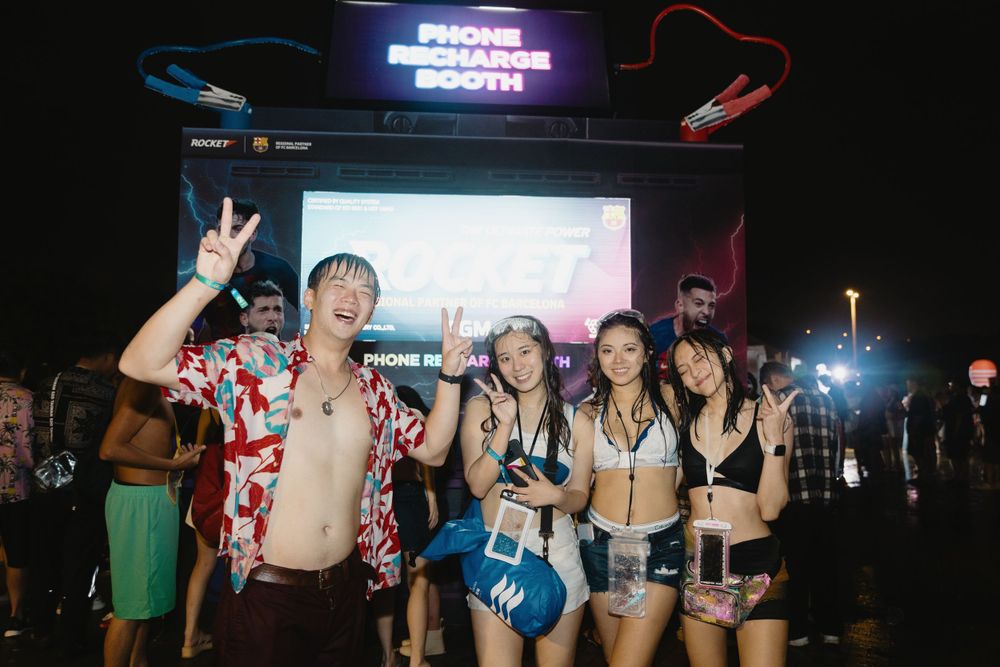 Group of young friends posing at a Phone Recharge Booth at a nighttime music festival.