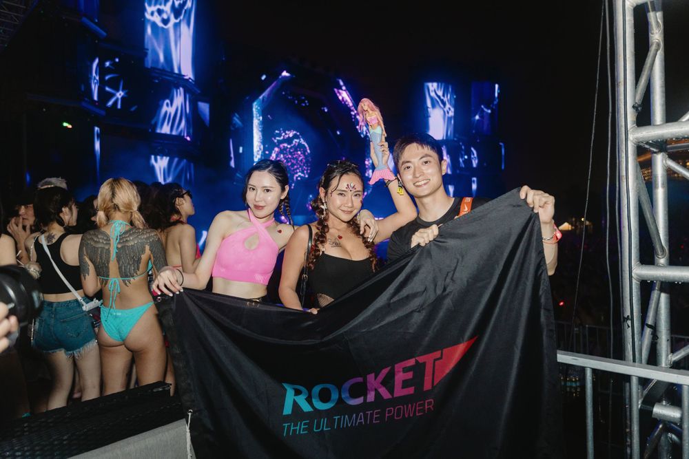 Three smiling women holding Rocket brand flag at energetic nightclub concert event.