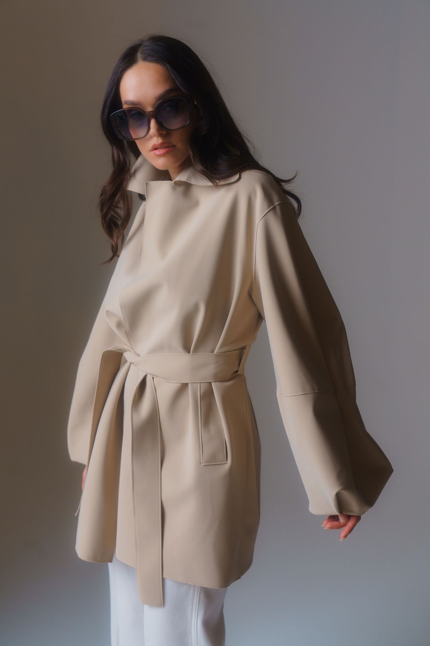 Elegant cream-colored belted coat styled with white pants and dark sunglasses in studio setting.