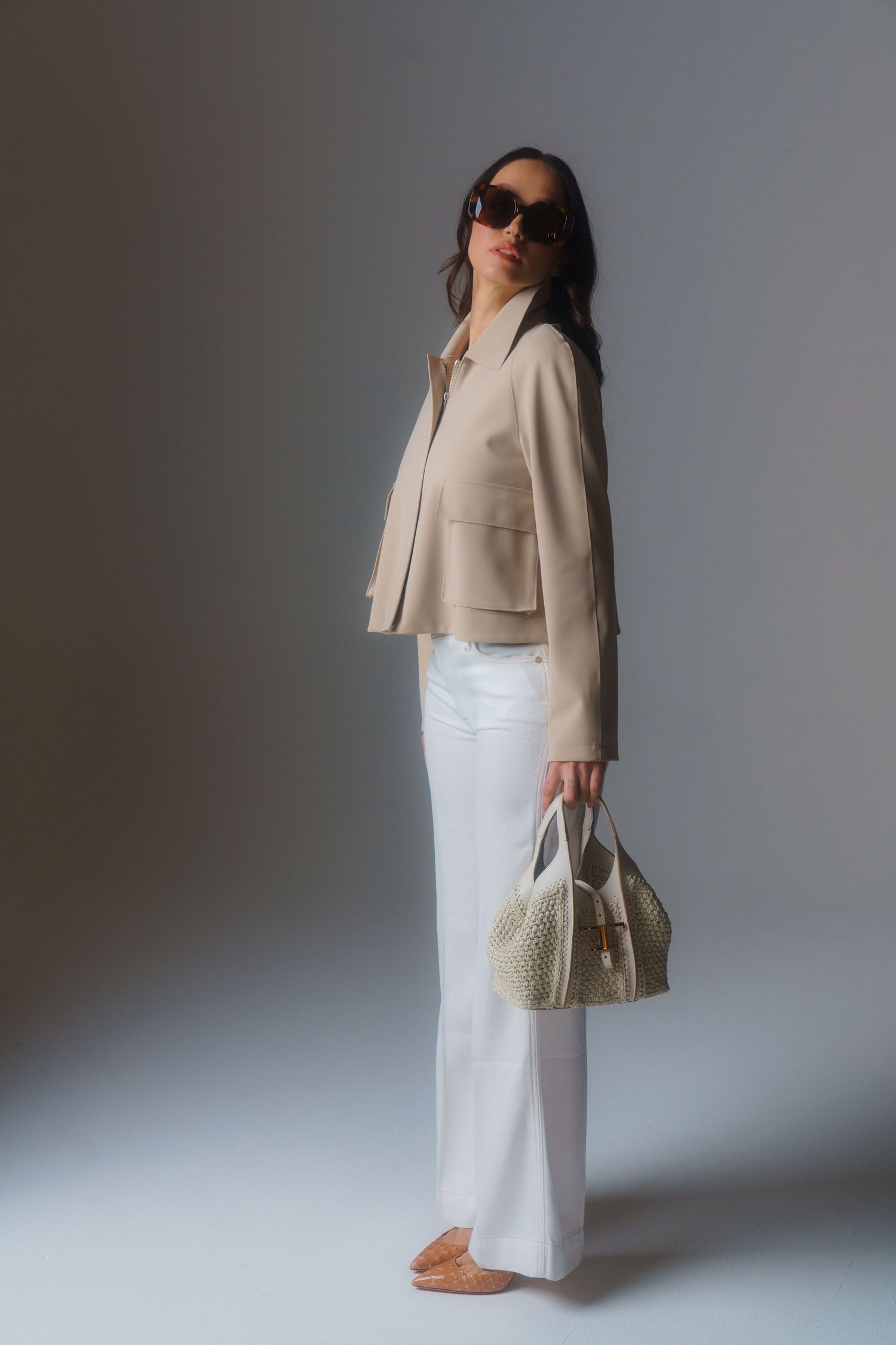 Minimal fashion look with beige jacket, white pants, and woven handbag against gray backdrop.